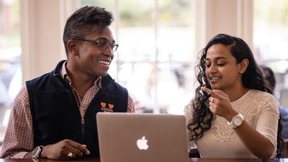 Two Darden students smiling and discussing something in front of a laptop.