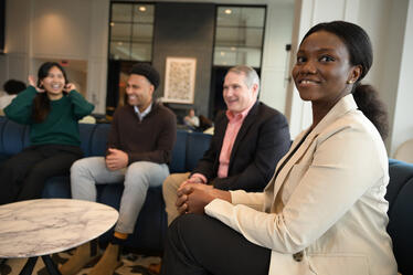Darden students and a faculty member sharing a lighthearted conversation in a lounge area, with one woman smiling in the foreground.